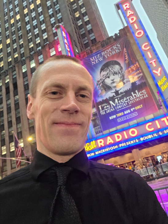A photo of me in a black dress shirt and a black tie, with a pattern on the tie, standing in front of Radio City Music Hall in New York City in August of 2026.