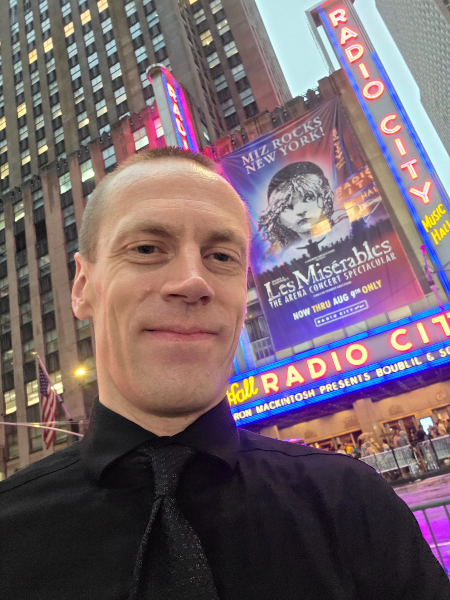 A picture of me in a black dress shirt and a black tie, with a pattern on the tie, standing in front of Radio City Music Hall in New York City in August of 2026.